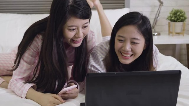 fast motion of two cheerful asian girl roommates laughing lying sitting on bed looking at laptop pc screen together talking chatting. young ladies in pajamas shopping online on christmas sale weekend