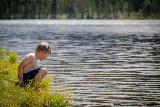 Summer nature view of a young caucasian boy in swimwear sitting in the grass looking in to the water.