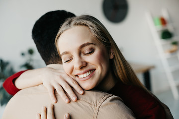 Happy positive young blonde woman hug guy. They stand in room. Model keep eyes closed. Blurred background