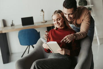 Young cheerful international couple in room. Latino guy stand behind woman and point. She sit in chair and read book. She hold it opened. They smile