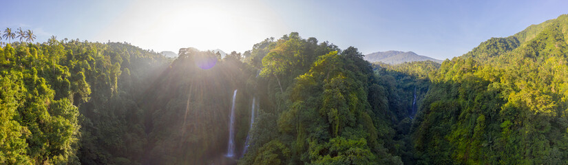 Aerial over Sekumpul waterfall surrounded by dense rainforest and mountains shrouded in mist at sunrise, Bali, Indonesia panoramic - obrazy, fototapety, plakaty