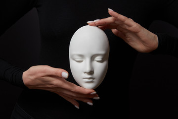 Two woman's hands hold white gypsum mask face on a black background. Concept social psychological masks - obrazy, fototapety, plakaty