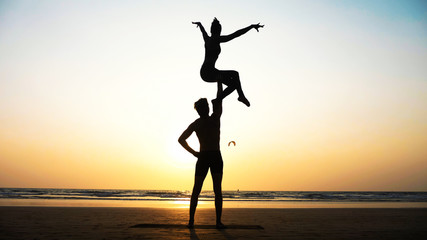 Fototapeta premium Silhouette of fit sporty couple practicing acrobatic yoga with partner together on the beach. Male standing and holding his female partner over his head. Mix race couple doing acrobatic exercise. 