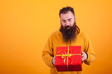 Smiling bearded man looking at the camera and holding a red gift box over yellow background.