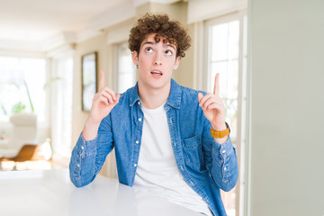 Young handsome man wearing casual denim jacket at home amazed and surprised looking up and pointing with fingers and raised arms.