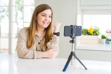 Beautiful young woman doing online video call using smartphone webcam smiling with happy face looking and pointing to the side with thumb up.