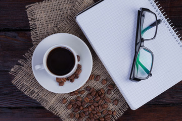 White coffee cup with notepad and glasses on a dark wooden background and linen fabric. Top view