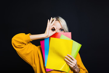 Cheerful blonde girl, hiding behind books, holding her fingers in the eye gesture of the eyepiece,...