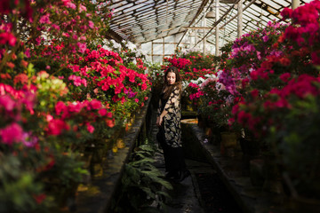 Young beautiful caucasian woman in glass greenhouse among colorful azalea flowers. Art portrait of a girl wearing a hat and romantic boho clothes. Gardening, plant growing, art therapy concept.