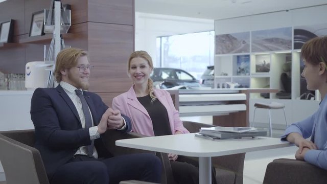 Saleswoman in a stylish suit transmitting the keys to the new car happy successful business couple. A joyful bearded guy in business suit happily picks up the keys of his new vehicle. Car showroom