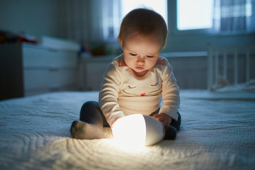 Adorable baby girl playing with bedside lamp in nursery