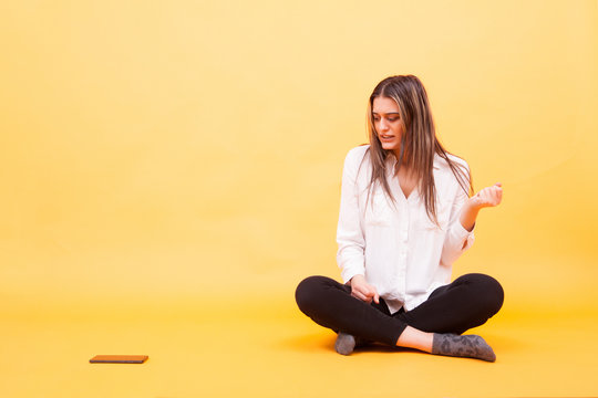 Beautiful young girl can't help herself not to loook at her phone while sitting down over yellow background