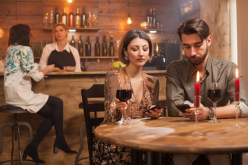 Beautiful wife and husband browsing on their phones while having a glass of red wine