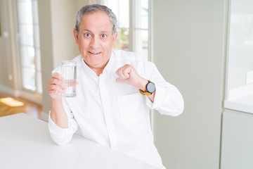 Handsome senior man drinking a fresh glass of water at home with surprise face pointing finger to himself