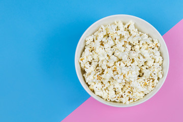 White bowl of popcorn on a bright pink and blue background with copy space. Movie/ film industry. Entertainment. Unhealthy food/ snacks.