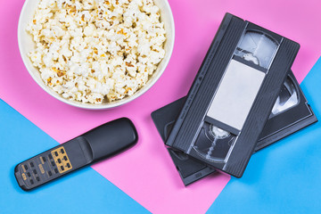 White bowl of popcorn, 2 VHS tapes and old fashioned remote control on a bright pink and blue background. Movie/ film industry. Entertainment. Unhealthy food/ snacks.