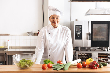 Photo of adult woman chef wearing white uniform cooking meal with fresh vegetables, in kitchen at...