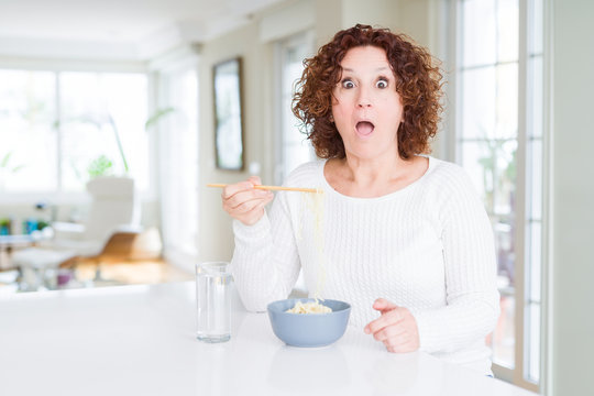 Senior woman eating asian noodles using chopsticks scared in shock with a surprise face, afraid and excited with fear expression