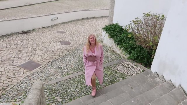 attractive blonde woman with long hair in a pink coat comes down slowly on the stairs, looks into the camera and smiles