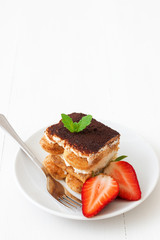 Tiramisu, homemade sweet Italian no bake dessert cheesecake, cut of cake on small plate embellished with fresh mint and a few real strawberries, on white table. Free space for your text. Closeup.