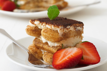 Tiramisu, homemade sweet Italian no bake dessert cheesecake, cut of cake on small plate embellished with fresh mint and a few real strawberries, on white table. Closeup.