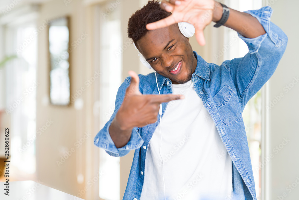 Poster African american man wearing headphones listening to music smiling making frame with hands and fingers with happy face. Creativity and photography concept.