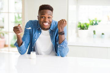 African american man eating healthy natural yogurt with a spoon screaming proud and celebrating...