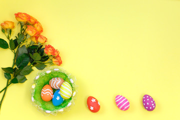 Beautiful fresh vivid orange roses tinged with red and colorful eggs in nest on yellow background.