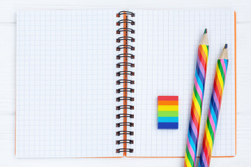 Open copybook with two rainbow-coloured pencils and a rubber on white wooden table