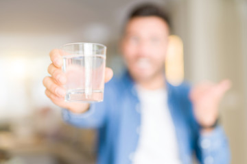 Young handsome man drinking a glass of water at home pointing and showing with thumb up to the side with happy face smiling