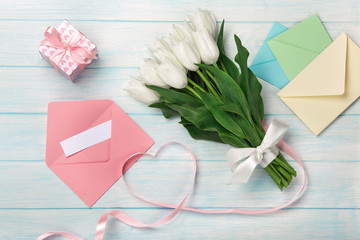 A bouquet of white tulips and a pink ribbon in the form of a heart with a gift box, love note and color envelope on blue wooden boards