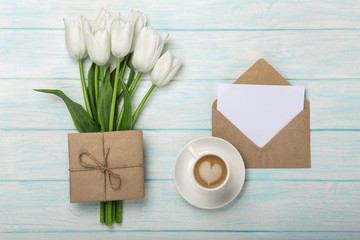 A bouquet of white tulips, cup of coffee with a love note and envelope on blue wooden boards