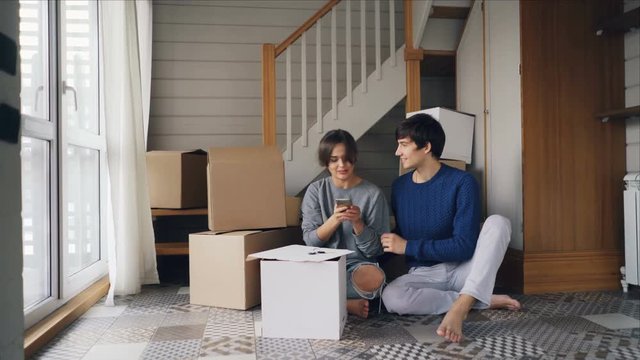 Happy couple is taking selfie after purchasing new apartment. Young people are posing and kissing looking at smartphone camera with boxes in background.