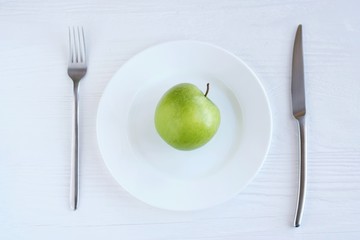 Juicy organic green apple on white plate with selective focus and blurred fork and steel knife on neutral wooden background. Diet concept with healthy vegetarian food for weight loss