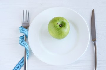 Juicy organic green apple on white plate with selective focus and blurred fork with blue measure tape on neutral wood background. Diet concept with healthy vegetarian food for weight loss