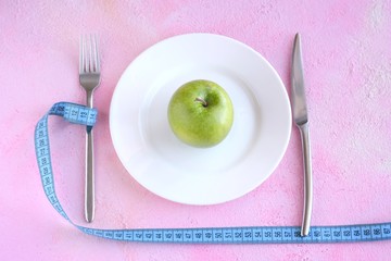 Juicy organic green apple on white plate with selective focus and blurred fork with blue measure tape on pink plaster background. Diet concept with healthy vegetarian food for weight loss