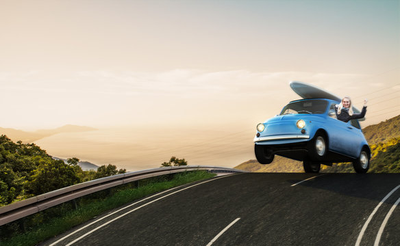 Kleines blaues Auto mit Surfbrett auf dem Dach rast &uuml;ber die Stra&szlig;e
