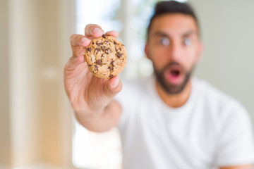 Handsome hispanic man eating chocolate chips cookies scared in shock with a surprise face, afraid and excited with fear expression