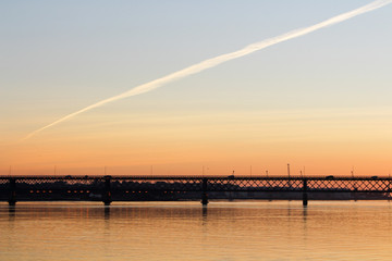 Obraz premium Bridge over the calm river with sunset colors. Plane trail in the sky