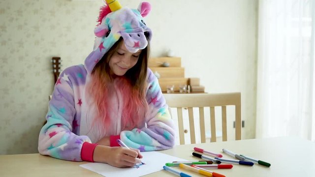 Cute happy teen girl wearing unicorn pajamas draws in notebook. homeschooling concept. Living rom in background