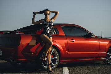 beautiful young woman with long dark hair in shorts sitting on the fender of red car on the sunset