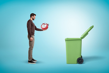 Man throwing broken red clock into open trash bin on blue background