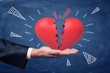 Side crop close-up of man's hand facing up and holding big red heart split into halves on blackboard background.