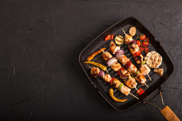 Grilled chicken skewers with spices and vegetables in a pan on black background