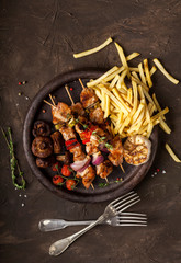 Grilled chicken skewers with spices, vegetables and fries in a pan on black background