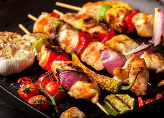 Grilled chicken skewers with spices and vegetables in a pan on black background