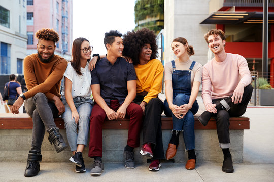 Six young adult friends sitting in a row on a bench in the street looking at each other, full length