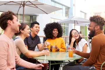 Six trendy millennial friends sit drinking cold brews talking and laughing outside a cafe
