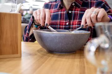 Woman in a plaid shirt holds a knife and fork in her hands with the help of which she is going to eat salad from a plate in a restaurant.