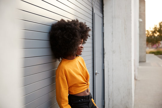 Young Black Woman With Afro Leaning In The Street Against Grey Security Shutters, Side View